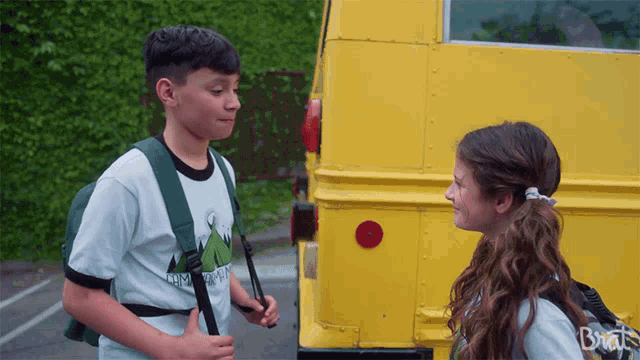 a boy and a girl are standing in front of a yellow bus with the word brat on the back