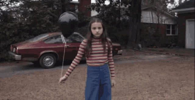 a girl in a striped shirt holds a black balloon in front of a red car