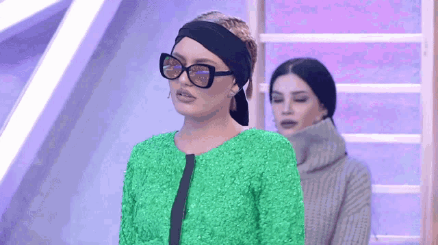 a woman in a green jacket and sunglasses stands next to another woman in a sweater