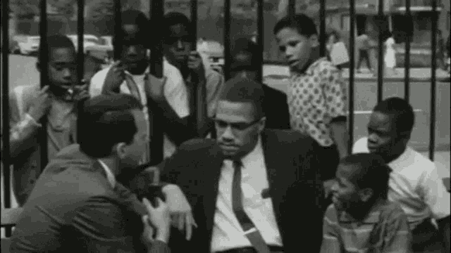 a black and white photo of a man in a suit and tie sitting in front of a fence surrounded by children .