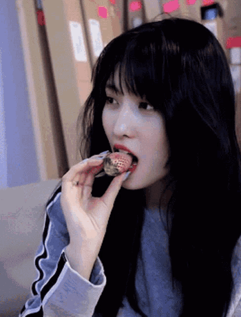 a woman with long black hair is eating a strawberry with her mouth open