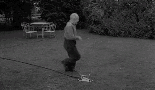 a black and white photo of a man walking on a lawn with a table and chairs in the background