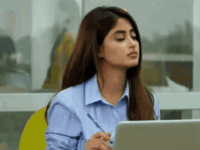 a woman in a blue shirt is sitting at a desk with a laptop and a pen in her hand .