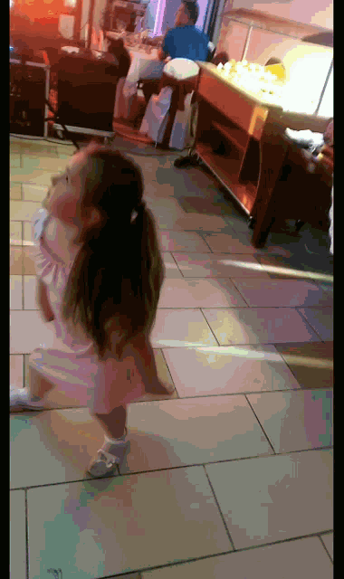 a little girl in a white dress and white shoes is walking on a tiled floor