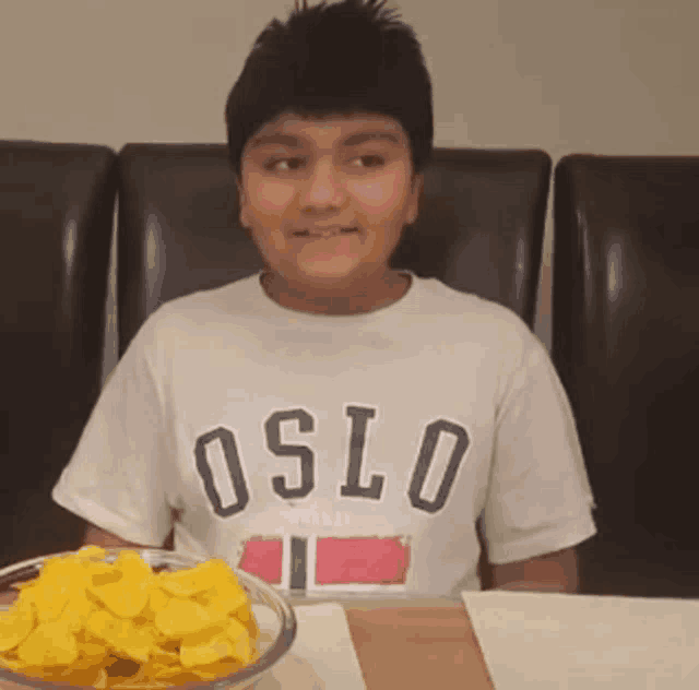 a boy wearing a white oslo shirt sits at a table with a bowl of chips