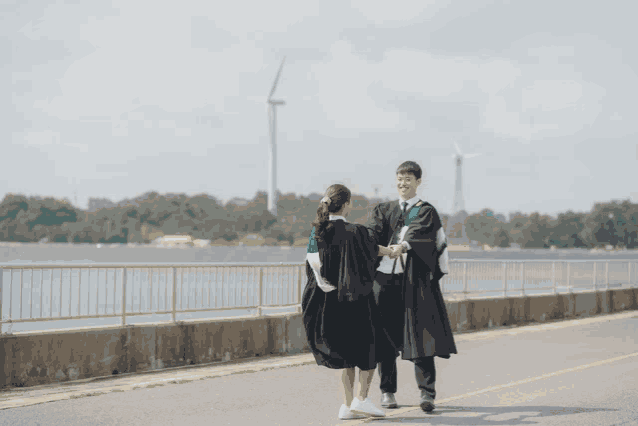 a man and a woman in graduation gowns are walking down a road with a windmill in the background