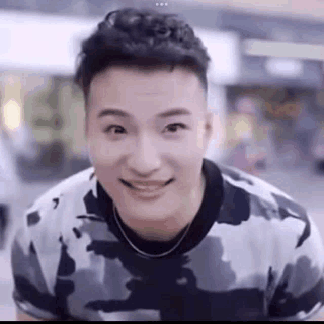 a close up of a man wearing a camouflage shirt and a necklace smiling .