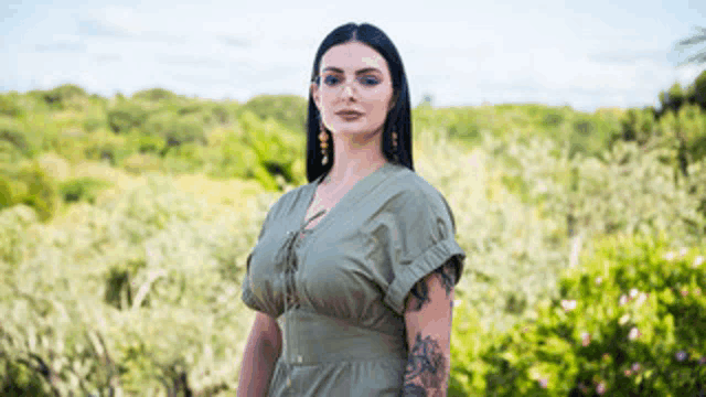 a woman in a green dress with a tattoo on her arm is standing in front of a forest .