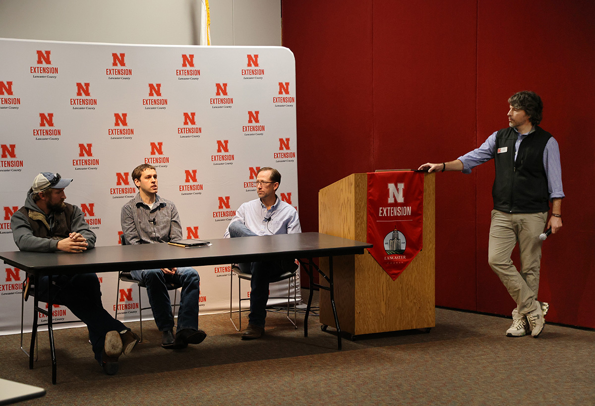 a person standing at a podium with a microphone, talking to three people sitting behind a table