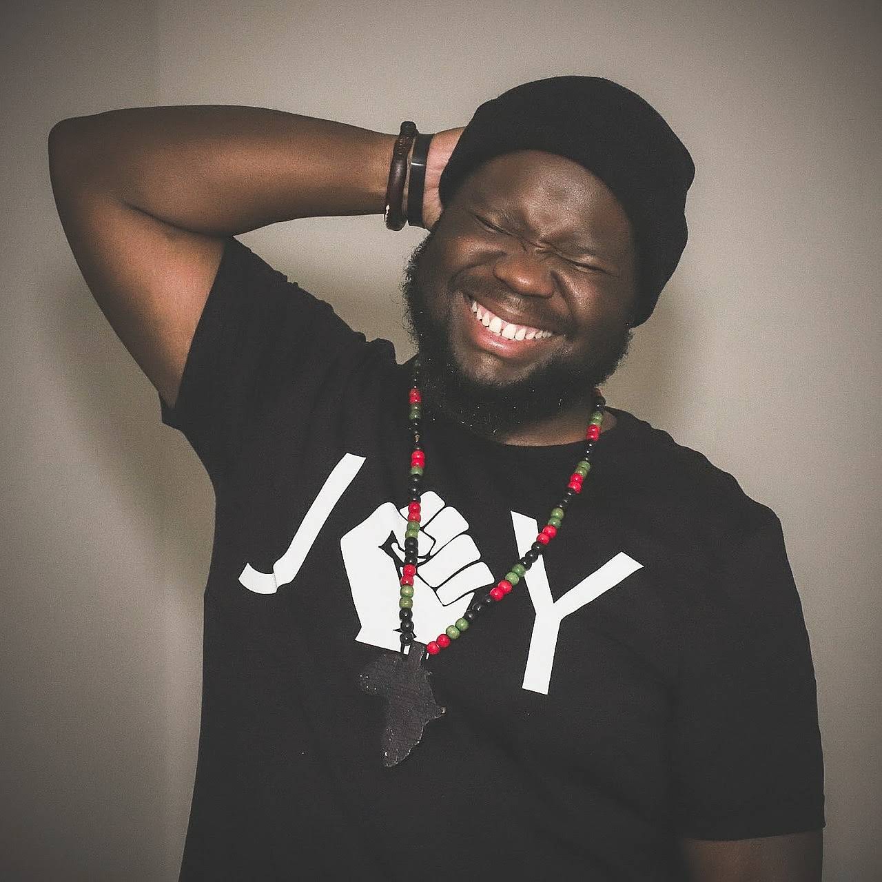 A person wearing a black beanie and a black shirt with the word JOY. They have a beard and are smiling with eyes closed, hand resting behind their head. A colorful beaded necklace with a large pendant hangs from their neck.