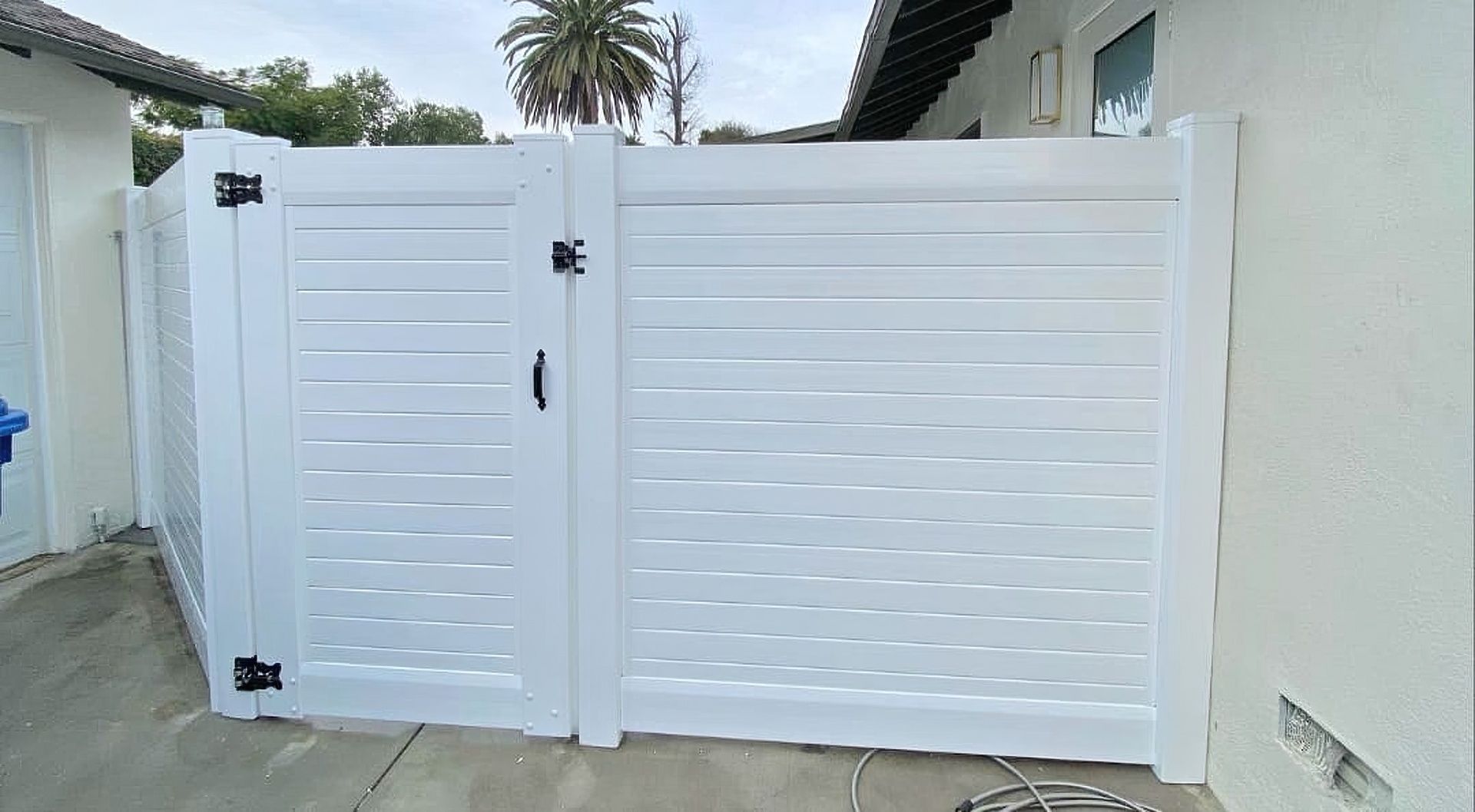 Vinyl fence gate opening to a suburban house with concrete floor and trees in the background.