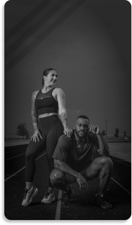 Two athletes pose on a running track at dusk; the woman is standing with hands on hips, the man is crouching, both looking confident.