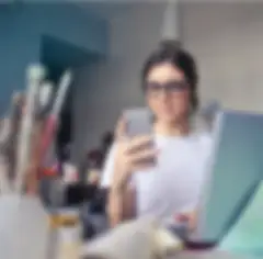 A young woman wearing glasses and a white t-shirt is looking at a cell phone while standing at an art table with cups of colored pencils and paintbrushes. There is a laptop open in front of her.