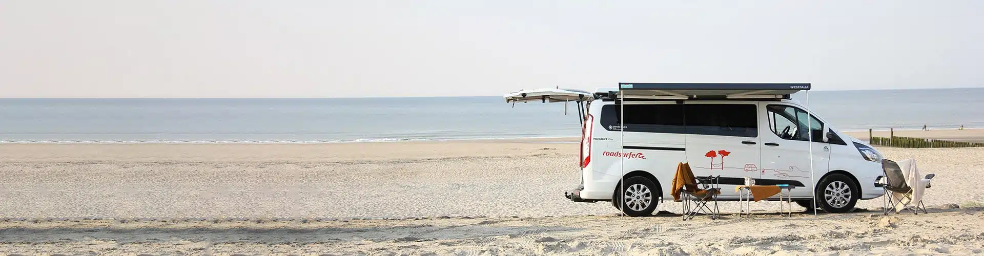 Camper Cabin Deluxe camper van from roadsurfer parked on an empty beach on a sunny day, awning open, table and chairs underneath