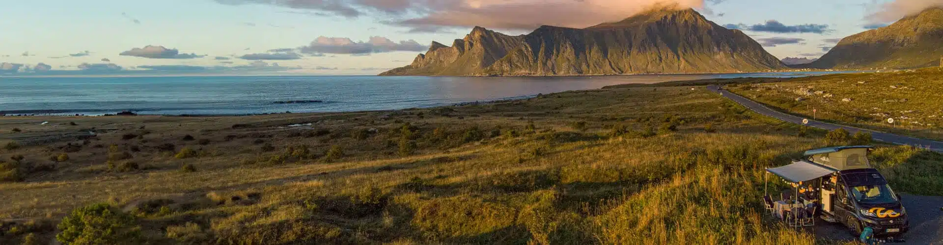 Campervan by the water next to green grass with hill panorama in the background at sunset in Norway