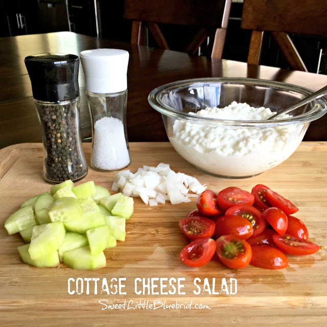 This photo shows cottage cheese in a clear glass mixing bowl with salt and pepper shakers, and onion, cucumber and tomato cut on a cutting board. 