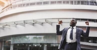 Happy excited black businessman celebrate victory with fists raised in the air staying against modern glass office center background, lifestyle portrait photo