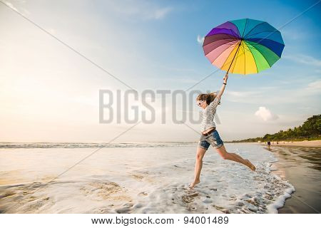 Cheerful young girl with rainbow umbrella having fun on the beach before sunset. Travel, holidays, v