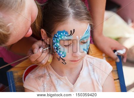 Child animator, artist's hand draws face painting to little girl. Child with funny face painting. Painter makes blue butterfly at girl's face. Children holiday, event, birthday party, entertainment.