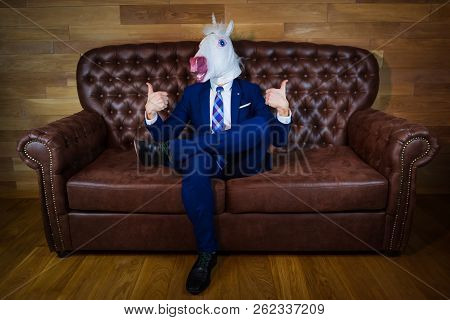 Funny Unicorn In Elegant Suit Sits On Sofa Like A Boss And Showing Gesture Thumbs Up. Portrait Of Un