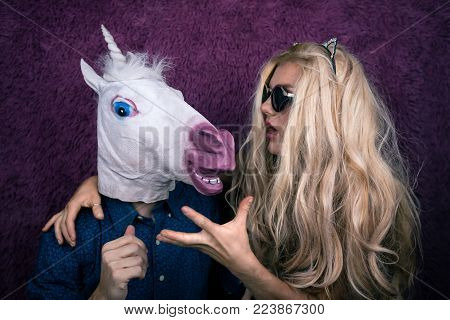 Portrait of happy unicorn in the arms of freaky young woman in sunglass and kitty ears on the purple background. Expressive blond with strange guy. Unusual people shows emotions.