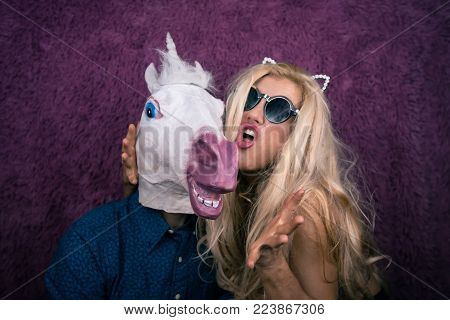 Portrait of happy unicorn in the arms of freaky young woman in sunglass and kitty ears on the purple background. Expressive blond with strange guy. Unusual people shows emotions.