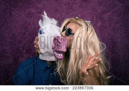 Freaky young woman in kitty ears kissing happy unicorn on the purple background. Expressive blond with strange guy. Unusual people sits together and shows emotions.