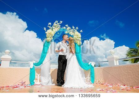 Wedding ceremony on a tropical beach in blue. Happy groom and bride under the arch decorated with flowers on the on hotel tropical sea in the background.