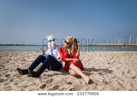 Young Unusual Man In Funny Mask Is Talking With Girlfriend. Elegant Attractive Travellers Sits On Th