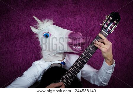 Freaky Young Man In Comical Mask Playing The Guitar On The Purple Background. Portrait Of Unusual Gu