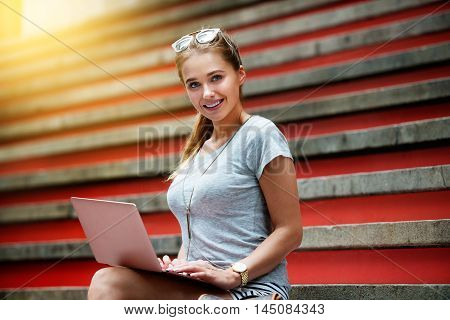 Beautiful student college girl sitting on steps and working on laptop computer. Happy woman with laptop outdoors.