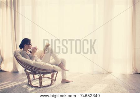 Young woman at home sitting on modern chair in front of window relaxing in her living room reading book and drinking coffee or tea