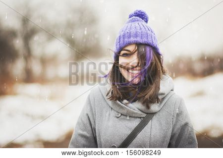 Portrait of cute, happy young woman in purple knitted beanie hat and gray coat, outdoors in winter on windy and snowy day, smiling and posing. Noise added for artistic effects.