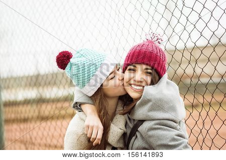 Two cheerful cute teenage girls hugging, one girl kissing her best female friend outdoors in winter. Two young women in knitted beanies and coats. No retouch.