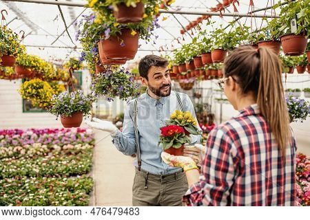 Young Smiling Gardener Chatting With His Female Colleague And Holding Pot With Yellow Flowers. Hotho