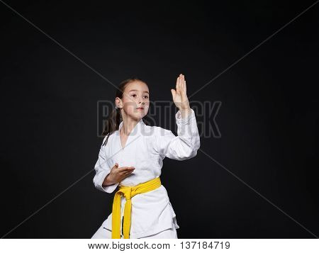 Little girl in karate suit kimono in studio at black background. Female child shows judo or karate stans in white uniform with yellow belt. Individual martial art sport for kids. Waist up portrait