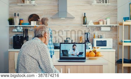 Senior Couple During Video Conference With Doctor Using Laptop In Kitchen Discussing About Health Pr