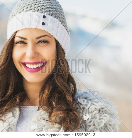 Closeup of gorgeous young brunette woman in knitted beanie hat in winter, posing, smiling, outdoors on snowy day in mountains. Closeup, copy space, retouched.