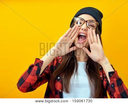 Hipster girl screaming like in megaphone holding hands near her face with open mouth. Young woman touts everyone over yellow background.