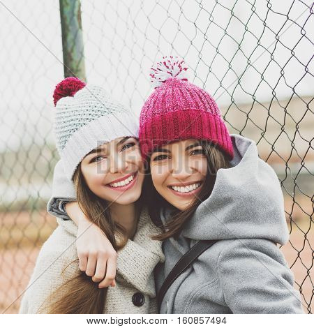 Two beautiful teenage girlfriends hugging and smiling outdoors in winter, wearing coats and knit beanie hats. Two young female friends portrait closeup. Retouched, natural light.