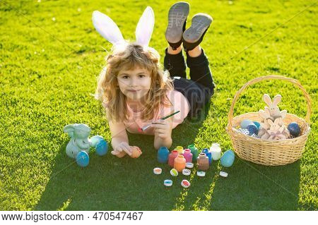 Child Laying On Grass In Park Wit Easter Eggs. Easter Kids Boy In Bunny Ears Painting Easter Eggs Ou