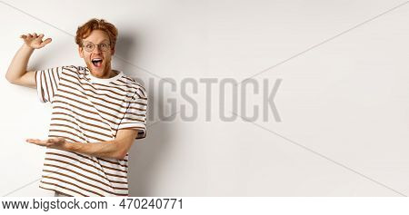 Amazed Redhead Man In Glasses Showing Something Large Size And Smiling At Camera, Saying Wow, Standi