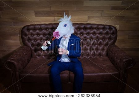 Funny Unicorn In Elegant Suit Sits On Sofa With Bottle Of Wine. Unusual Man With Headache At Home. F