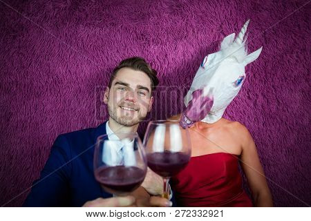 Portrait Of Strange Couple On Purple Background. Freaky Young Woman In Comical Mask Drinking Wine Wi