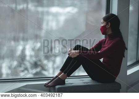 Winter depression because of coronavirus confinement. Sad Asian woman alone during city lockdown wearing face mask indoors at home covid. Anxiety, stress, mental health issues.