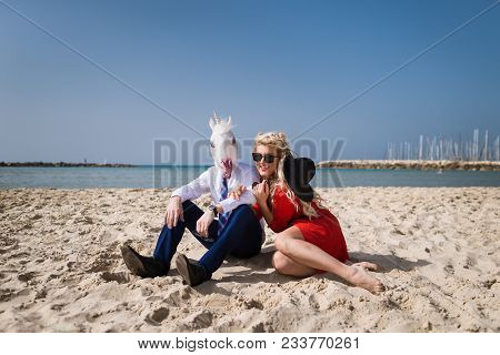 Freaky Guy In Suit And Funny Mask Sits With Beautiful Girl In Red Dress On The Beach. Young Happy Wo