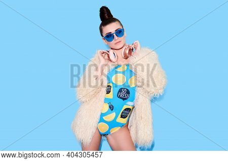 Freaky girl dj with pink headphones wearing swimsuit and fur coat over blue isolated background