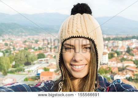 Selfie of young woman in knitted beige modern beanie hat. Closeup self portrait of beautiful blonde teenage girl, town urban area in the background. Natural light, mild retouch.