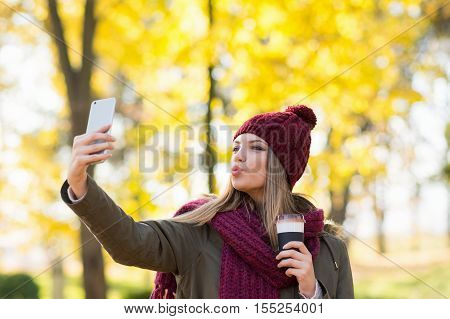 Young woman with takeaway coffee taking a selfie on smart phone in park in autumn. Millennial teenage girl in fall taking a photo of herself using cellphone. Vibrant colors, natural light.