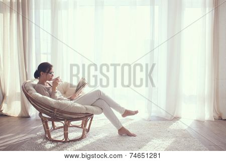 Young woman at home sitting on modern chair in front of window relaxing in her living room reading book and drinking coffee or tea